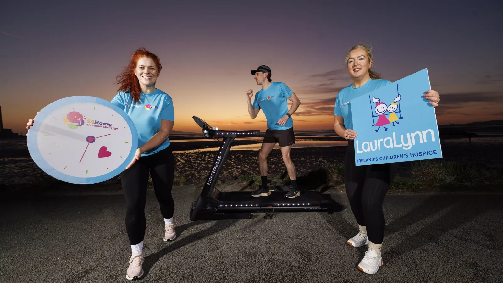 Sunrise at Sandymount Strand. There is a person running on a treadmill. Two other people are holding boards that say 24 hour treadmill challenege and LauraLynn.
